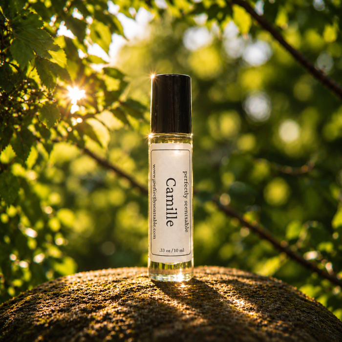 Camille roll-on perfume bottle displayed on a mossy stone outdoors, surrounded by lush green leaves with warm sunlight filtering through the trees in the background.