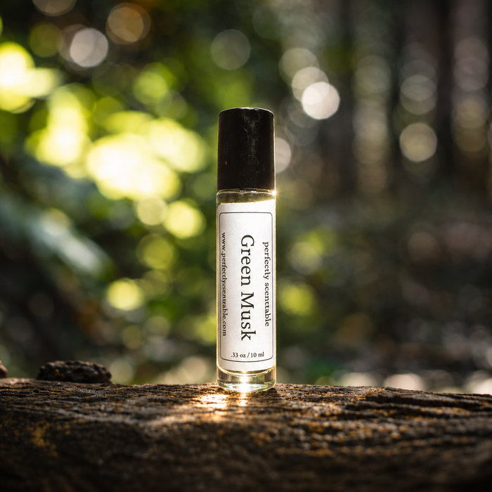 Green Musk roll-on perfume bottle standing on a natural wooden surface, with a softly blurred green forest background and warm sunlight creating glowing bokeh highlights.