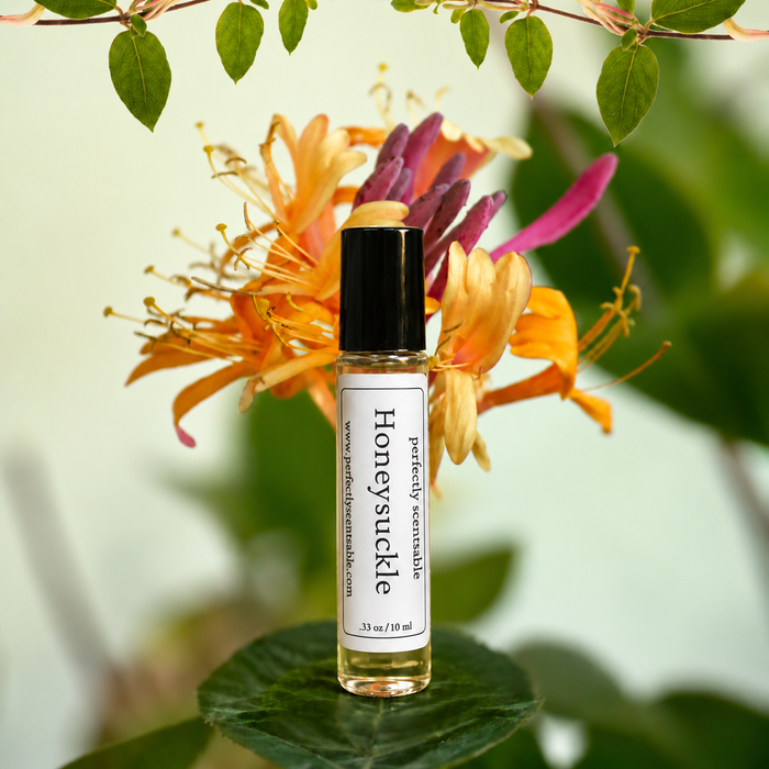 Honeysuckle roll-on perfume bottle displayed on a green leaf in front of vibrant orange, yellow, and pink honeysuckle flowers, surrounded by fresh green foliage in soft natural light.