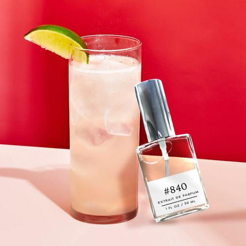Bottle of Perfectly Scentsable #840 Extrait de Parfum leaning beside a tall iced cocktail with a lime wedge, set against a bright red background.