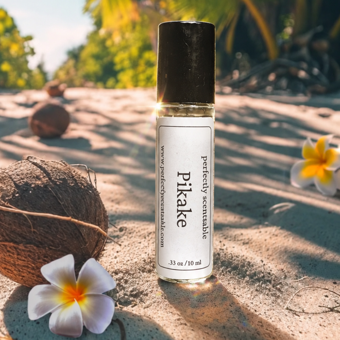 Pikake roll-on perfume bottle standing on a tropical sandy beach beside a coconut and white plumeria flowers, with palm trees and warm sunlight in the background.