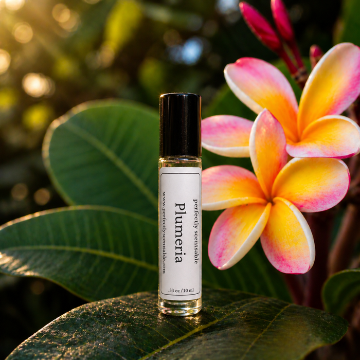 Plumeria roll-on perfume bottle displayed on a glossy green leaf beside vibrant pink, orange, and yellow plumeria flowers, with warm natural light and soft greenery in the background.