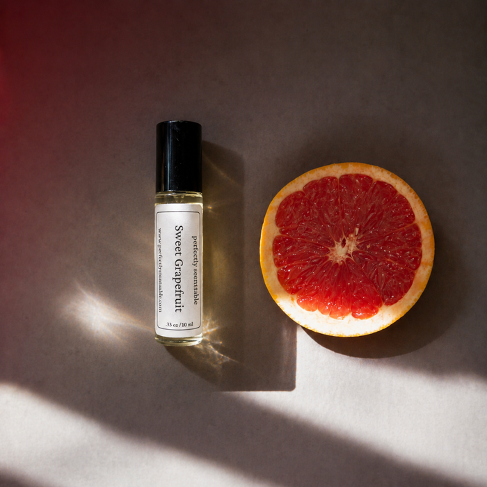 Sweet Grapefruit roll-on perfume bottle displayed beside a halved ruby-red grapefruit, illuminated by warm directional light that casts soft shadows and glowing reflections across a neutral surface.