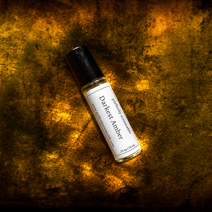 Darkest Amber roll-on perfume bottle lying diagonally on a richly textured golden-brown surface, illuminated by warm amber light and dramatic shadow patterns.