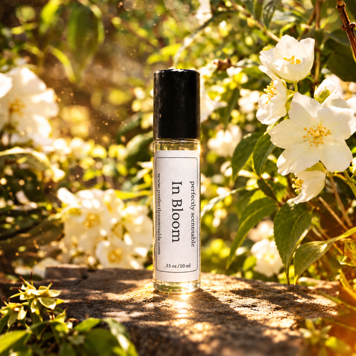 In Bloom roll-on perfume bottle displayed on a sunlit stone surface, surrounded by white blossoms and lush green leaves with warm golden light filtering through the garden.