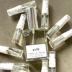Clear bottle labeled with #199 Extrait De Parfum with silver cap, accompanied by 9 elegantly arranged sample bottles, resting on a marble surface.