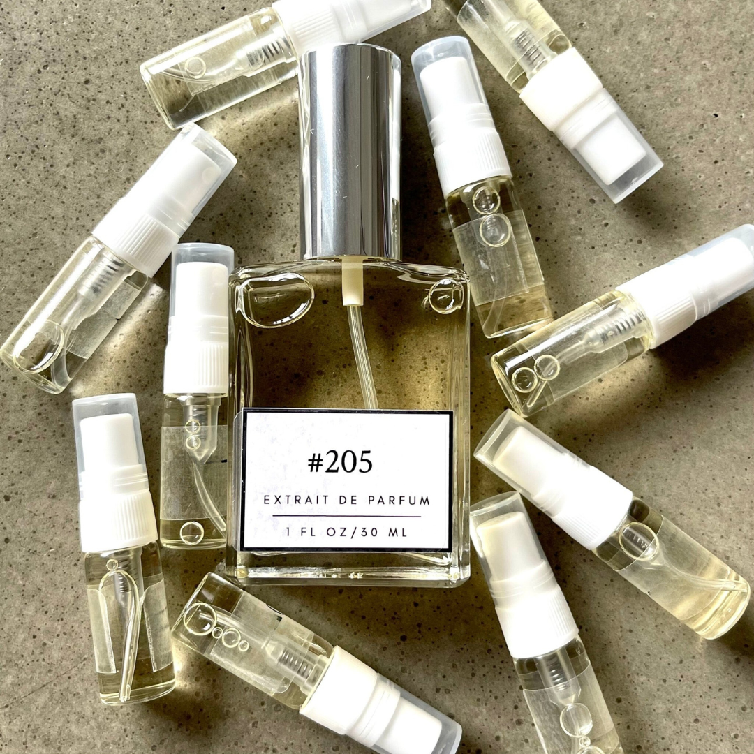 Clear bottle labeled with #205 Extrait De Parfum with silver cap, accompanied by 10 elegantly arranged sample bottles, resting on a marble surface.