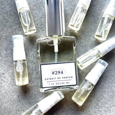 Clear bottle labeled with #294 Extrait De Parfum with silver cap, accompanied by 7 elegantly arranged sample bottles, resting on a marble surface.