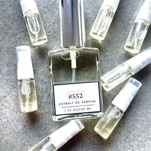 Clear bottle labeled with #552 Extrait De Parfum with silver cap, accompanied by 7 elegantly arranged sample bottles, resting on a marble surface.