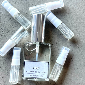 Clear bottle labeled with #567 Extrait De Parfum with silver cap, accompanied by 6 elegantly arranged sample bottles, resting on a marble surface.