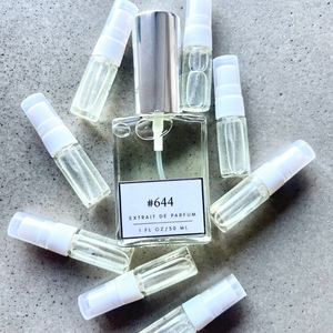 Clear bottle labeled with #644 Extrait De Parfum with silver cap, accompanied by 8 elegantly arranged sample bottles, resting on a marble surface.