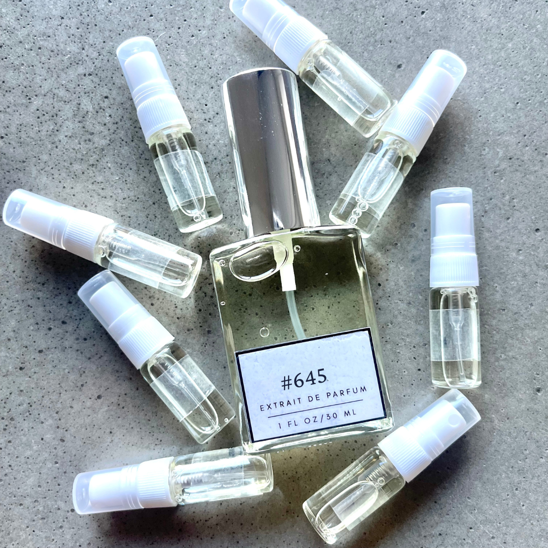 Clear bottle labeled with #645 Extrait De Parfum with silver cap, accompanied by 8 elegantly arranged sample bottles, resting on a marble surface.
