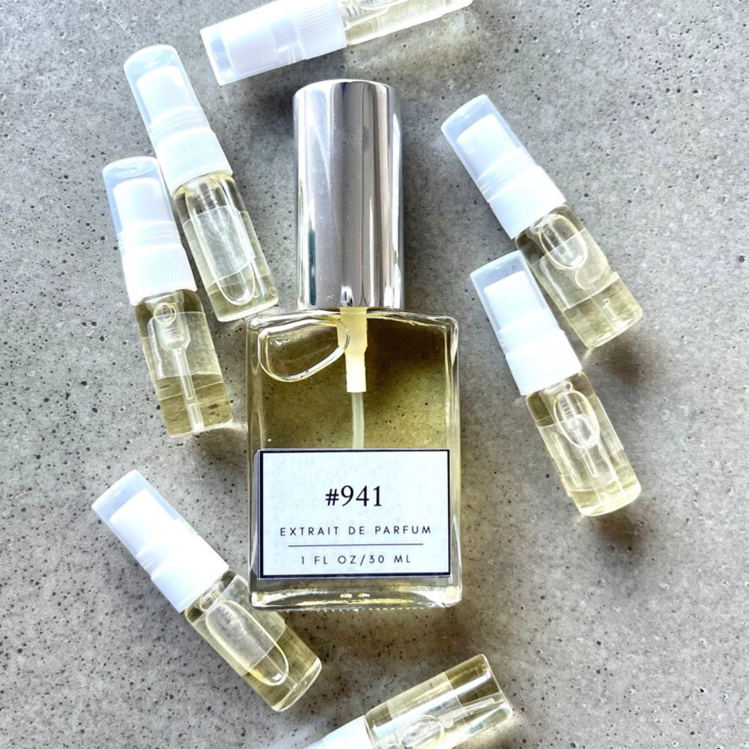 Clear bottle labeled with #941 Extrait De Parfum with silver cap, accompanied by 7 elegantly arranged sample bottles, resting on a marble surface.