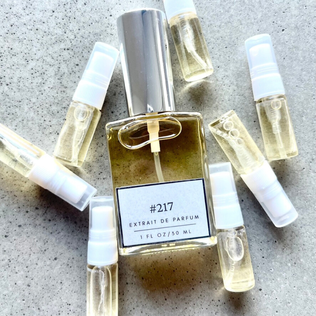 Clear bottle labeled with #217 Extrait De Parfum with silver cap, accompanied by 7 elegantly arranged sample bottles, resting on a marble surface