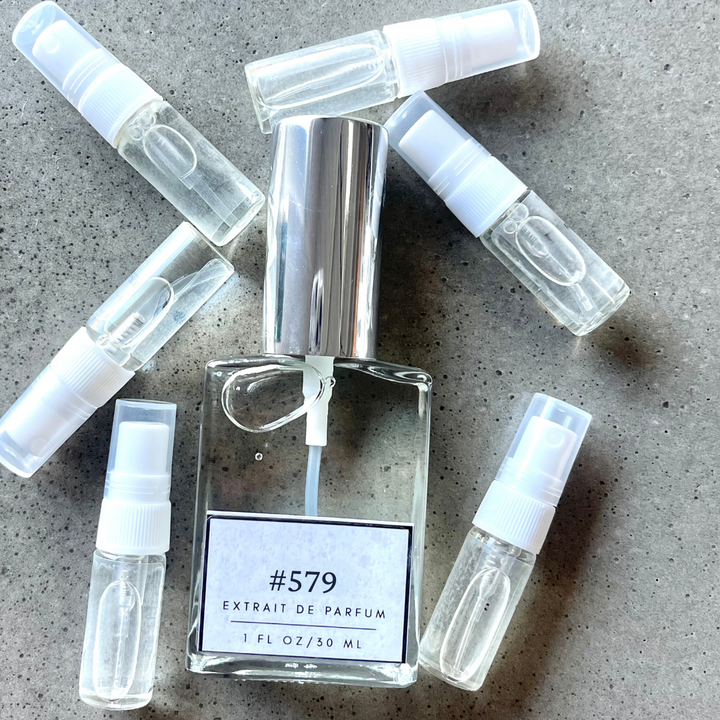 Clear bottle labeled with #579 Extrait De Parfum with silver cap, accompanied by 6 elegantly arranged sample bottles, resting on a marble surface