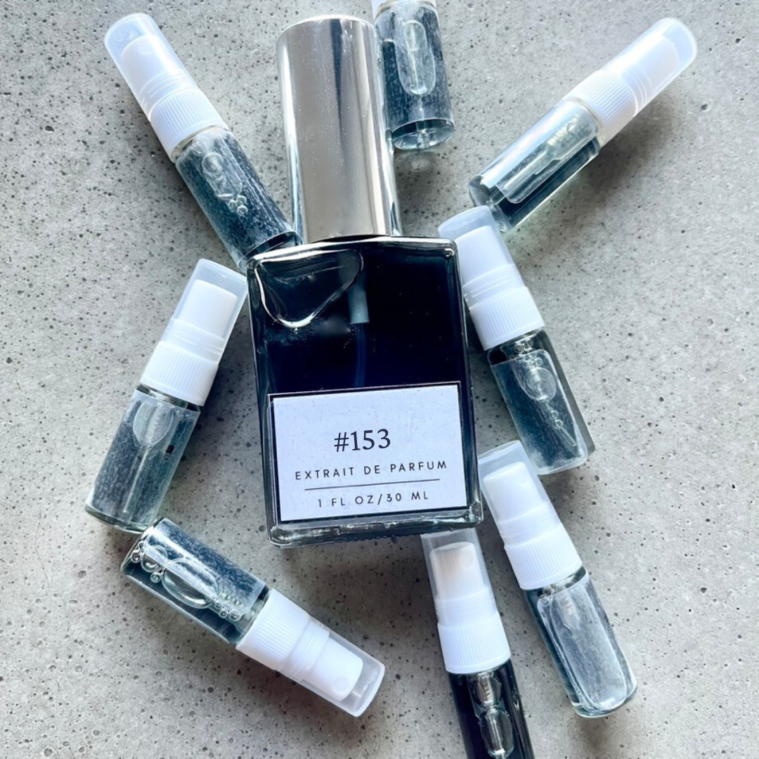 Clear bottle labeled with #153 Extrait De Parfum with silver cap, accompanied by 8 elegantly arranged sample bottles, resting on a marble surface.