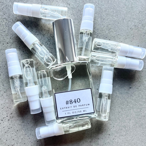 Clear bottle labeled with #840 Extrait De Parfum with silver cap, accompanied by 10 elegantly arranged sample bottles, resting on a marble surface.