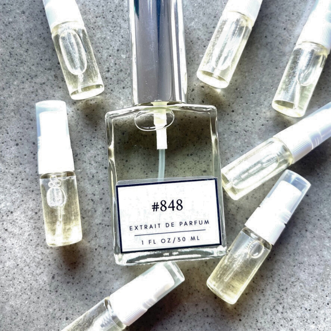 Clear bottle labeled with #848 Extrait De Parfum with silver cap, accompanied by 7 elegantly arranged sample bottles, resting on a marble surface.