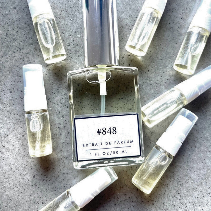 Clear bottle labeled with #848 Extrait De Parfum with silver cap, accompanied by 7 elegantly arranged sample bottles, resting on a marble surface.