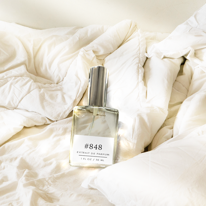 Bottle of fragrance dupe 848 Dupe Lazy Sunday Morning Inspiration' with a floral scent, labeled '#848 extrait de parfum 1 fl oz/30 ml', lying on white sheets bathed in gentle sun rays.