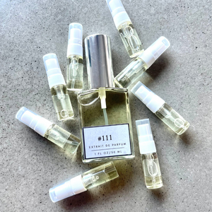 Clear bottle labeled with #111 Extrait De Parfum with silver cap, accompanied by 8 elegantly arranged sample bottles, resting on a marble surface