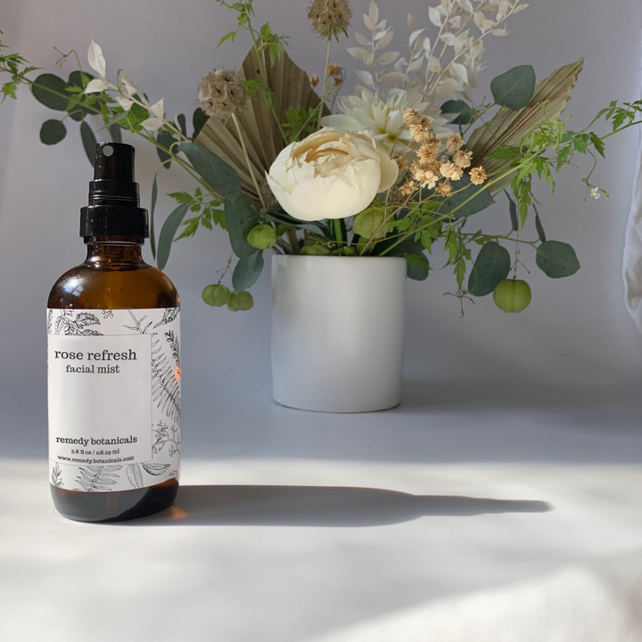 "A bottle of Rose Refresh Facial Mist stands on the left side, centered next to a variety of flowers arranged in a white vase, set against a soft gray background. The arrangement evokes freshness and natural beauty.