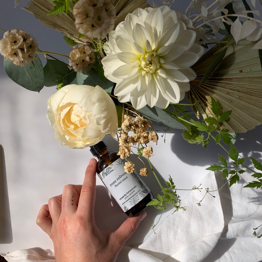 A rose refresh facial mist bottle laying  on a table beside a top view depth of a variant flower.